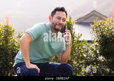 Mature Asian man sitting on low wall in residential garden, holding smartphone and talking Stock Photo