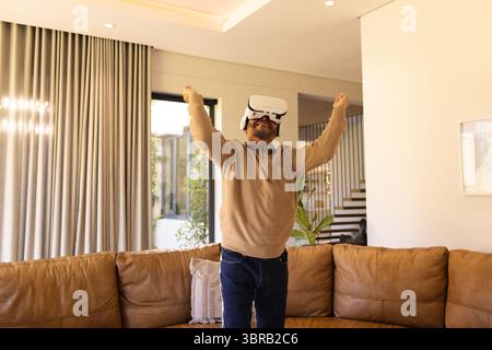 Senior Asian man raising arms while wearing white VR headset in living room near caramel sofa Stock Photo