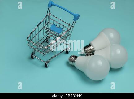 Three LED light bulbs and shopping cart on blue background. Stock Photo