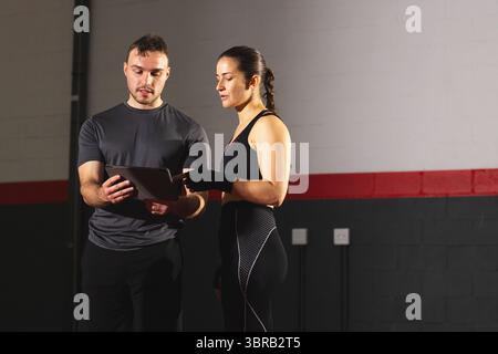 Male fitness trainer and female client standing together reviewing workout plan on tablet in gym Stock Photo