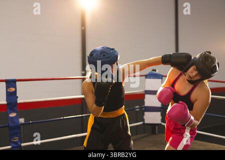 Two female boxers sparring in gym ring under bright lights wearing gloves and headgear, copy space Stock Photo