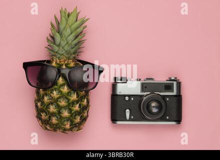 Summer background. Fun and humor. Travel concept. Pineapple with sunglasses retro camera on a pink pastel background. Top view Stock Photo