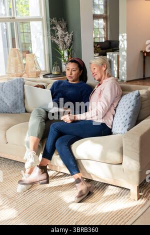 Female friends sitting on beige sofa using laptop in living room beside textured pillows Stock Photo
