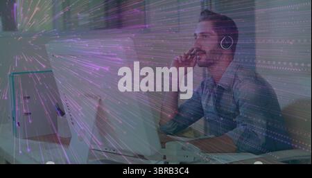 IT pro wearing blue shirt thinking in office with two monitors, keyboard, handset, mug, headphones Stock Photo