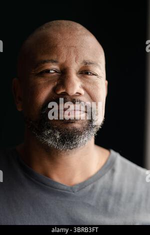 Senior African American man smiling and posing in room with dark backdrop and window light Stock Photo
