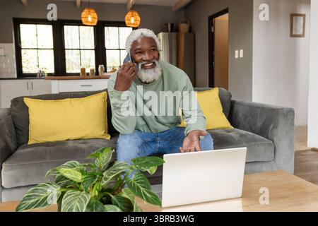 Senior African American man calling on smartphone sitting at home on sofa with laptop on table Stock Photo