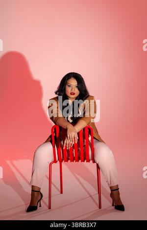 Asian woman posing backwards on red metal chair in studio, wearing tan blazer and pink trousers Stock Photo
