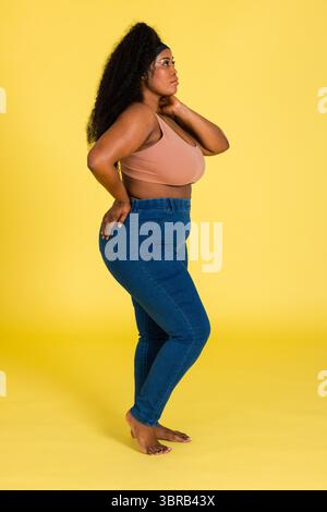 African American woman modeling in peach sports bra and blue skinny jeans in yellow studio backdrop Stock Photo
