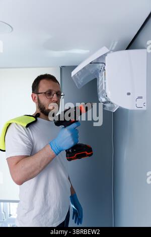 Man Utilizing a Power Drill for Installing a Wall-Mounted Air Conditioning Unit in a Modern Indoor Space with Safety Gloves and a Towel on His Stock Photo