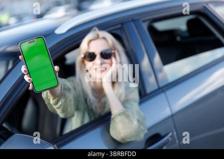 A Cheerful Young Woman Showcasing Her Smartphone with a Bright Green Screen While Relaxing Inside Her Car, Enjoying a Perfect Day Out Stock Photo