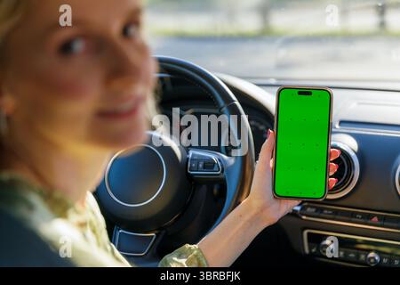 Smiling Woman in Car Displaying Smartphone with Green Screen While Driving, Perfect for Template Use in Advertisements and Digital Content Creation Stock Photo