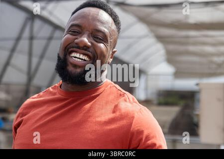 African American man standing in greenhouse framed by metal beams under shade cloth among planters Stock Photo