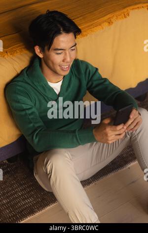 Asian man sitting on patterned rug against sofa with yellow throw holding smartphone in living room Stock Photo