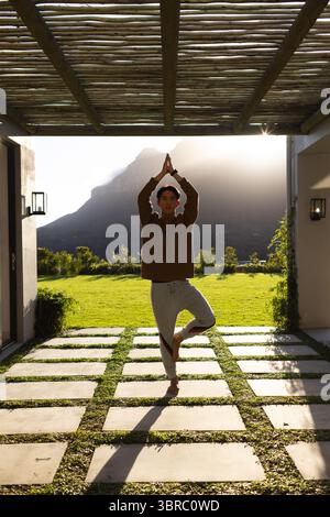 Asian man practicing tree pose on stone patio under pergola overlooking mountain view at sunrise Stock Photo