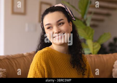 Woman sitting on couch in living room wearing mustard sweater, pink scarf with framed art, plant Stock Photo