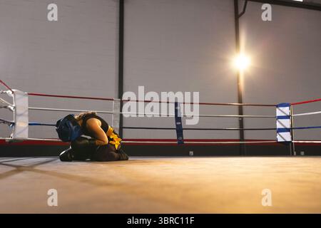 female boxer kneeling on ring mat under spotlight wearing blue headgear and gloves, copy space Stock Photo