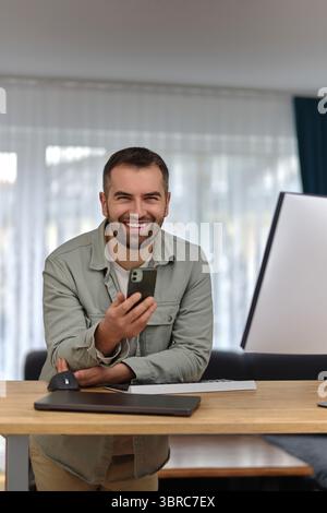 A young happy man using his phone mobile while working at home office, looking at the camera and smiling. Stock Photo