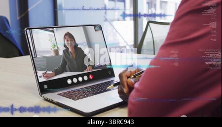 Sitting man in burgundy shirt joining call on laptop in glass-walled office, with pen, notebook Stock Photo