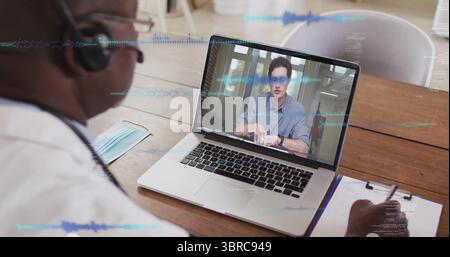Doctor in white coat taking notes during call in office, with laptop, headset, mask and clipboard Stock Photo