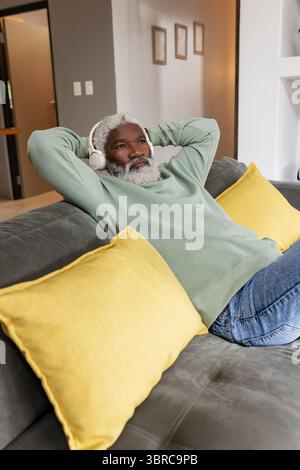 Senior African American man reclining on grey couch in living room listening with white headphones Stock Photo