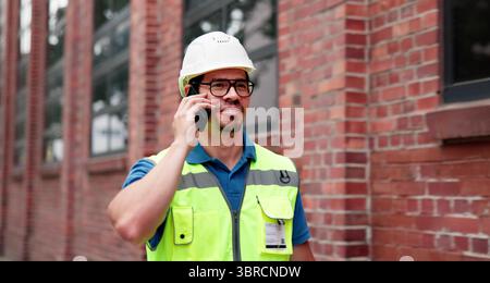 Skilled Builder In Uniform Discussing Progress Over Phone Call Efficiently. Stock Photo