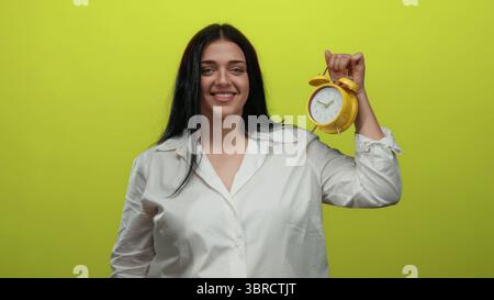 Young woman in white shirt holding yellow clock smiling against isolated yellow background Stock Photo