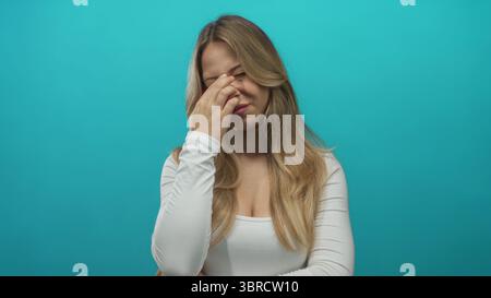 Young caucasian blonde woman pinches nose bridge under studio lights; stress headache fatigue tension. Stock Photo