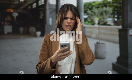 Hispanic woman holding smartphone looks distressed while standing outdoors in busy city street, showing signs of a headache under the daylight. Stock Photo