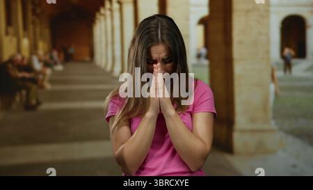 Woman in pink shirt praying with eyes closed in ancient university courtyard. Stock Photo