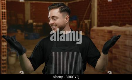 Young man with beard wearing apron and gloves smiling at construction site, suggesting indoor service or work in progress. Stock Photo