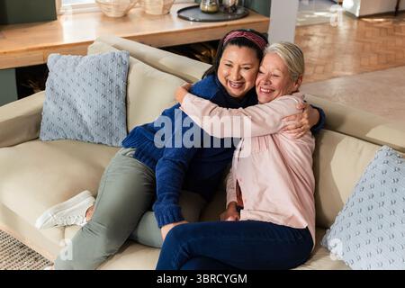 Diverse senior friends sitting on sofa at home hugging near throw pillows and console table Stock Photo