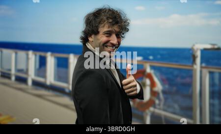 Handsome man smiling confidently on a cruise ship outdoors with the ocean in the background, wearing a suit and giving a thumbs up Stock Photo