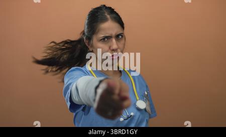 Young hispanic woman doctor with stethoscope beckons with outstretched hand in studio; invitation command. Stock Photo