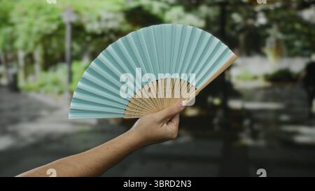 Man holding blue fan outdoors in park setting on sunny day showcasing traditional accessory with blurred green background. Stock Photo