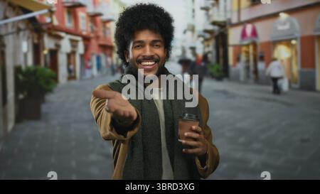 Smiling man with afro holding coffee on city street wearing scarf and jacket, reaching out happily amid urban surroundings with vibrant buildings and Stock Photo