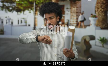 African american man holding blueprints on city street checks time looking at smartwatch. Stock Photo