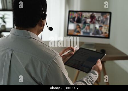 A virtual meeting and virtual conference person participating virtual meeting using headset, tablet, and computer monitor displaying multiple people Stock Photo