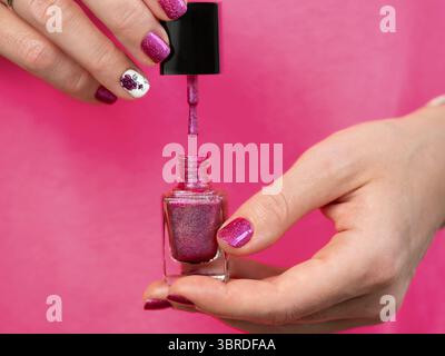 Female hands with pink manicure has pink purple nail polish bottle. Woman holding pink holographic nail polish lacquer brush. Aesthetic visual of red Stock Photo