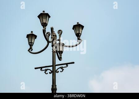 An ornate vintage-style black iron street lamp with four classic lanterns against a clear light blue sky. Decorative, old-fashioned urban lighting. Stock Photo