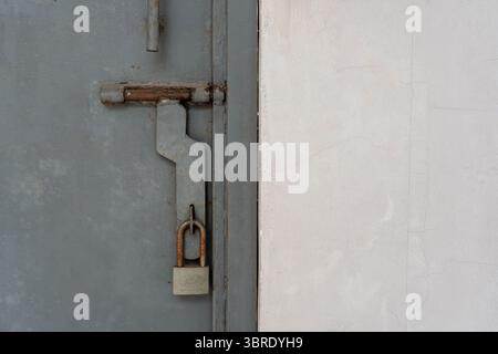 Close-up of a rusty old padlock securing a slide bolt on a gray industrial metal door. Concept of security, privacy, or being locked away. Stock Photo