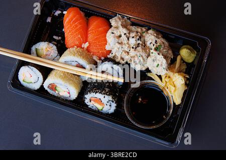 A takeaway sushi meal in a black plastic tray with salmon nigiri, maki rolls, ginger, wasabi, and soy sauce with chopsticks over the tray. Selective f Stock Photo