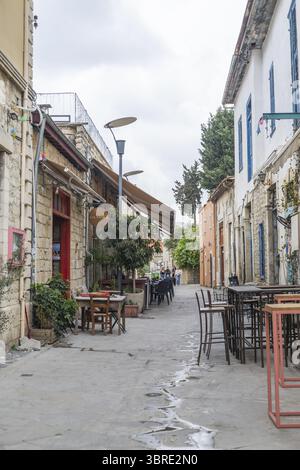 Limassol, Cyprus - 25 June 2025: View of a narrow street lined with tables and chairs outside quaint buildings, casting shadows under an overcast sky. Stock Photo