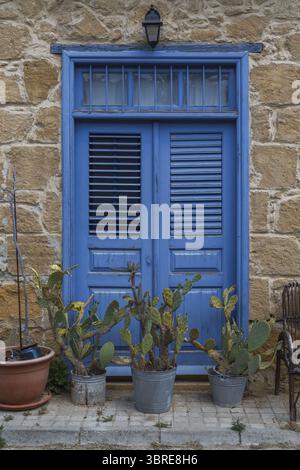 Lefkosia, Cyprus - 25 June 2025: View of a vibrant blue door set against a textured stone wall, framed by prickly pear cacti and a vintage lantern. Stock Photo