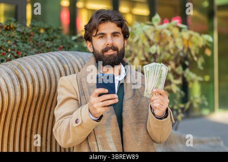 Happy rich Indian businessman tourist using smartphone and counting money dollar cash banknotes sitting on bench on downtown city street. Success busi Stock Photo