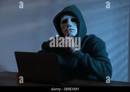 Anonymity. Masked man with laptop showing hush gesture at table in dark Stock Photo