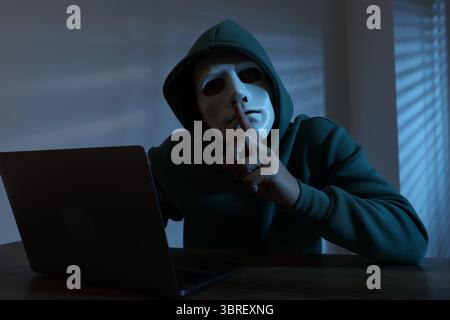 Anonymity. Masked man with laptop showing hush gesture at table in dark Stock Photo