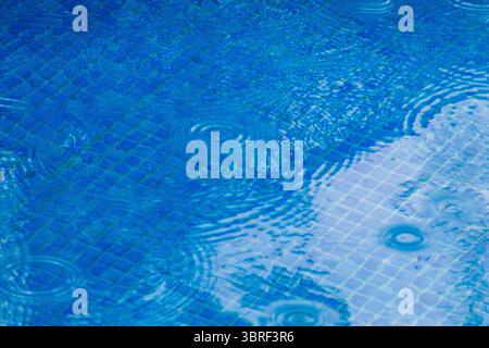 Raindrops splashing against the serene surface of a blue swimming pool, creating mesmerizing ripples on a warm summer day Stock Photo