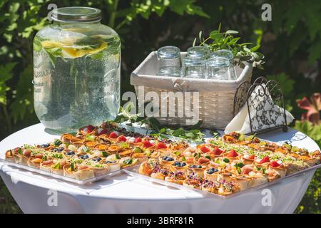 Table covered with a white tablecloth with a jar of fresh water, a basket with glasses, napkins and canapes or starter, during an outdoor party Stock Photo