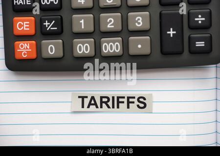 A grey strip of paper with the word Tariffs typed on it lying in an open notebook near a calculator. Stock Photo
