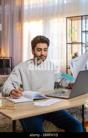 Young Indian man freelancer sitting at table reviewing financial reports on laptop, holding documents and receipts, taking notes in notebook. Hispanic guy sits nearby at home, focused on his own task. Stock Photo